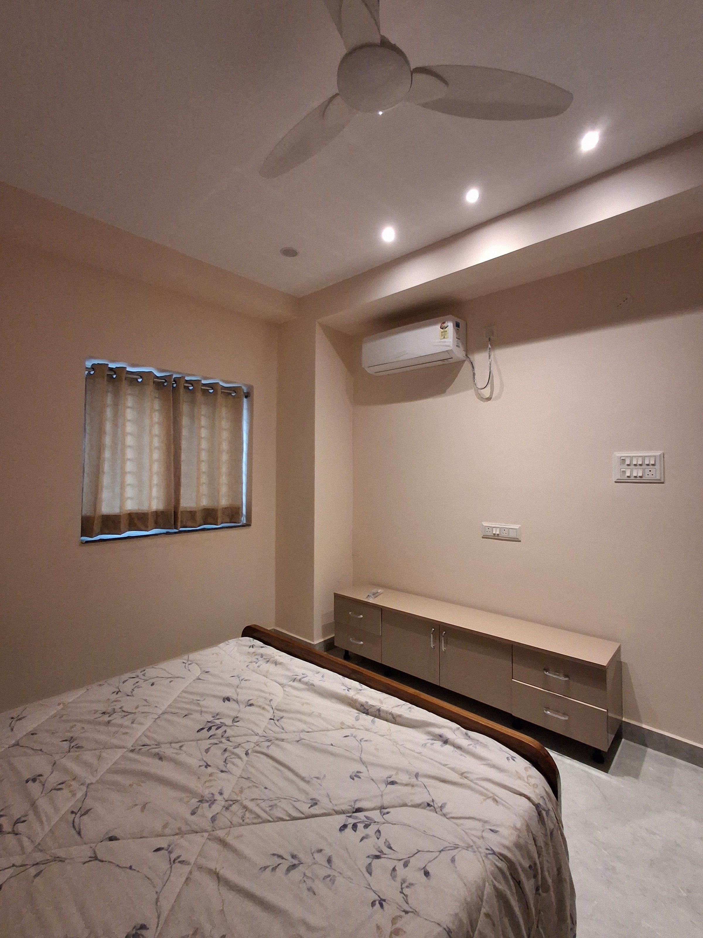A bedroom photographed in portrait and tilted from beside the bed: the foot of the bed in the lower left corner, a curtained window on the left wall, a split AC and a low dresser on the far wall, under a heavy band of ceiling, in a pinkish evening cast.