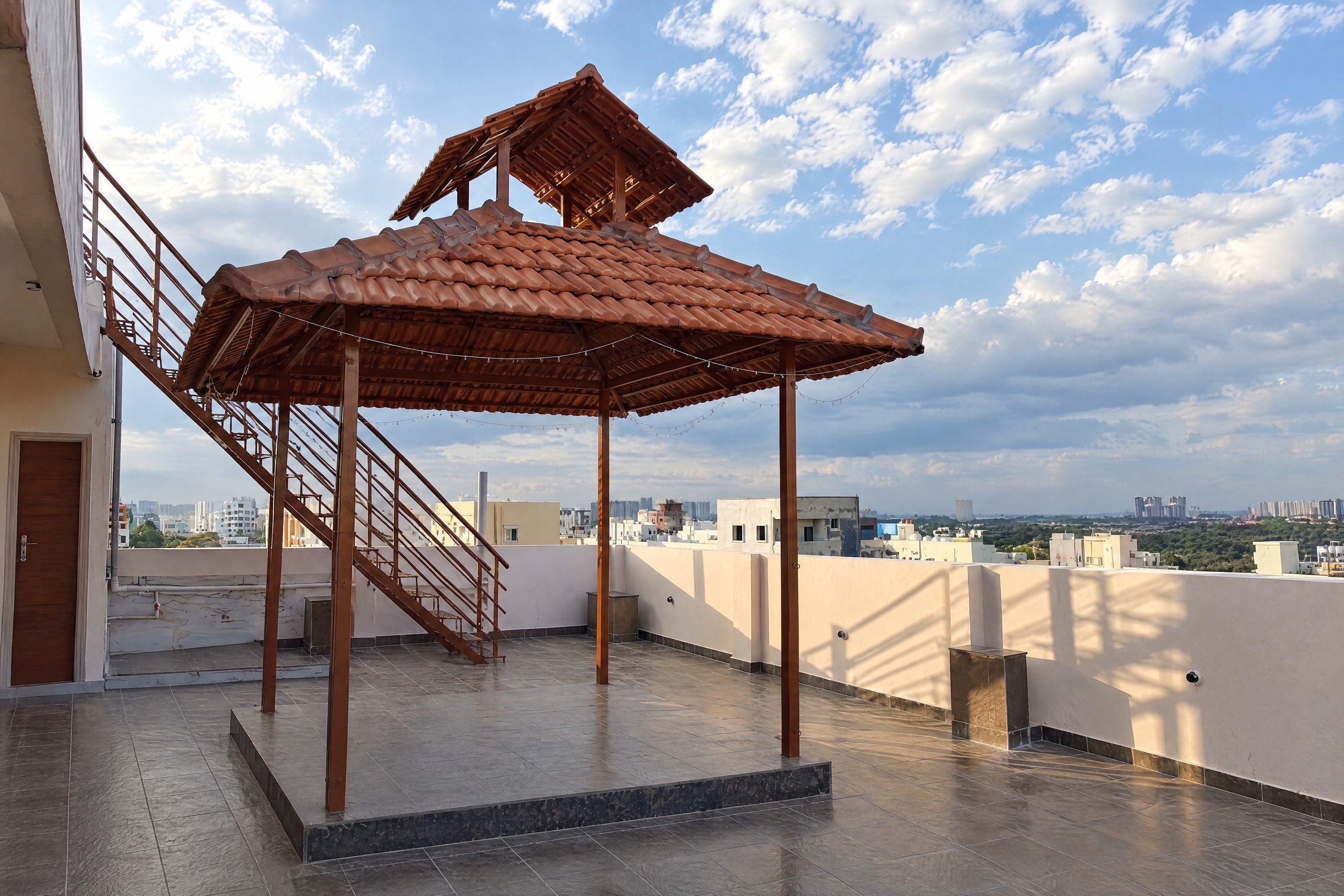 An AI re-angle of a terrace as a level landscape frame: the gazebo on its raised platform, the staircase climbing behind it on the left to the upper deck, the flat’s door at the left edge and the length of the parapet, under a brighter sky.